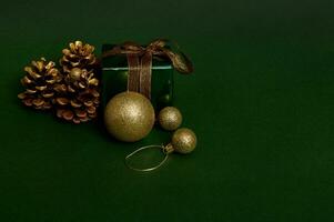 Luxury present in shiny green glitter wrapping gift paper with gold ribbon and golden bow, pines cones and spherical Christmas tree toys laid out on dark background. Copy space for advertisement photo