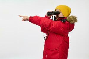 Curious school boy, adventurer, explorer in warm bright clothes looking through binoculars and pointing with finger on a copy space of a white background. photo