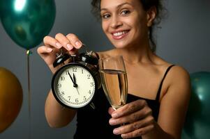 Close-up of an alarm clock and flute of champagne in the hands of blurred smiling beautiful woman, isolated over gray background and inflated green metallic and golden air balloons, copy space for ad photo