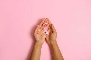 Top view of hands holding pink ribbon on pink background, Breast cancer awareness, October Pink day, World Wancer Cay, national Cancer Survivor Say. Women's health care and medical concept. Copy space photo