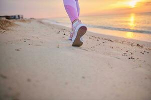 Closeup of runner legs in pink sports shoes performing jog on a sandy beach during sunrise in the early morning photo