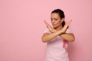 Confident portrait of beautiful serene African American woman wearing pink t-shirt and cancer awareness ribbon, gesturing STOP with hand, looking at camera posing over pink background with copy space photo