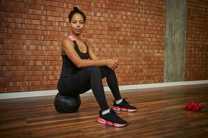 Beautiful African athlete woman in stylish black activewear sitting on a medicine ball and looking at camera at gym studio photo