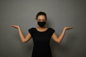 Isolated waist length portrait on gray background of confident serene African woman wearing black protective medical mask raising hands palms up while holding imaginary copy space for advertisement photo