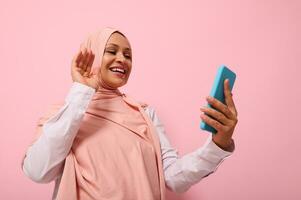 musulmán linda mujer de medio oriental o árabe etnia con cubierto cabeza en rosado hijab teniendo un vídeo conferencia en móvil teléfono, saludo su interlocutor durante vídeo llamar, rosado antecedentes Copiar espacio foto