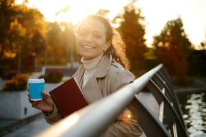 Happy cheerful young woman with a blue takeaway paper mug of coffee and hand book in the autumnal park, smiles looking at camera with falling sunbeams on the background of yellow golden leaves. Autumn photo