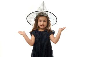 Isolated portrait of pretty little witch girl wearing wizard hat, dressed in stylish carnival dress, gesturing, holding a copy space imaginary on the palm to insert an advertising. Halloween concept photo