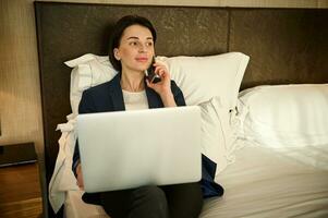 Attractive smiling young Caucasian business woman relaxing on a bed at hotel bedchamber, working on laptop computer, talking on mobile phone during a business trip photo