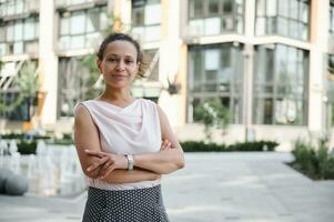 Confident business portrait of a successful middle aged business woman in casual attire posing to camera with crossed rams on the background of high buildings. Urban city backdrop photo