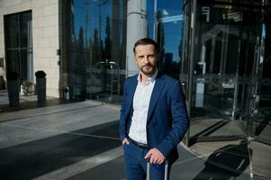 Handsome businessman developer entrepreneur wearing business suit looks at camera posing with a suitcase and his hand in pocket on the background of corporate modern high-rise building glass entrance photo