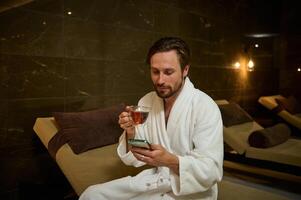 Handsome attractive Caucasian man in bathrobe relaxing, using mobile phone, browsing web sites and drinking herbal tea, sitting on a chaise lounge by jacuzzi in luxurious Ayurvedic wellness spa resort photo