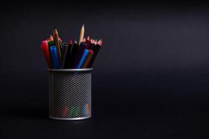 Stand for pens and pencils on the desktop on a black background with copy space. Close-up of a metal bucket with stationery. Holder with colored school supplies photo