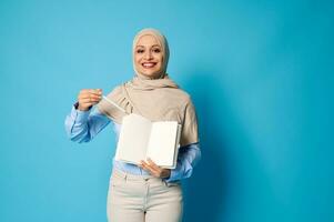 Charming Muslim woman in hijab with a beautiful smile points a pen on a white blank sheet of paper on a daily photo