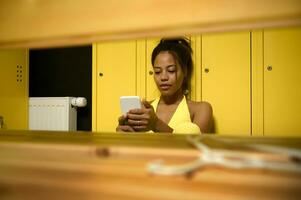 View through a wooden bench to female African athlete, sportswoman in yellow tight-fitting tracksuit, resting after heavy workout, sitting on the floor in gym locker room, swiping in smartphone photo