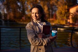 Lifestyle portrait of pretty woman in warm beige coat holding takeaway cup of coffee and hugging her book in hardcover, smiling on the autumn park and lake background. Leisure activity, happy weekend photo