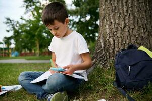 Elementary aged schoolboy concentrated, focused on solving school tasks, sitting next to a tree on green grass and holding a mobile phone, making notes, writing on workbook. Back to school concepts photo