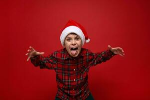 Funny cheerfule cute European preadolescent boy in plaid shirt and Santa Claus hat grimacing looking at camera posing against red colored background with copy space for New Year and Christmas ad photo