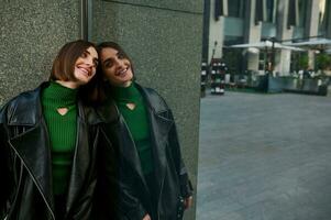 Fashionable cheerful pretty woman in eco-leather clothes posing for camera, leaning against the marble wall of a modern building, smiles toothy smile looking at her reflection on the glass entrance photo