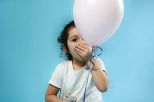 adorable niño niña cubre su boca con su mano y poses a cámara detrás un rosado globo en azul antecedentes con Copiar espacio foto