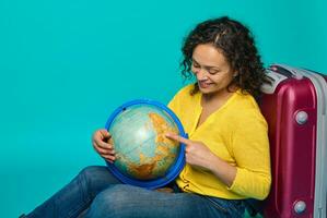 Charming woman traveler in yellow shirt sits next to a suitcase and points with her finger on a place on the globe, choosing a travel destination. Tourism concept on blue background with copy ad space photo