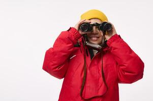 Adorable school boy in yellow woolen hat and bright red down jacket, smiles toothy smile looking through binoculars, isolated on white background with copy space. Winter travel and adventure concept photo