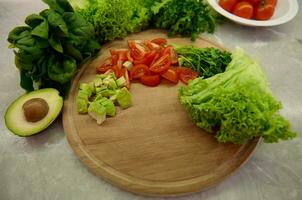 High angle view of chopped tomatoes and avocado on a wooden board, variety of greens, lettuce, salad and spinach leaves on a kitchen countertop. Healthy ingredients for raw vegan salad photo