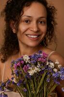 Close-up portrait of charming smiling woman holding a bouquet of beautiful wildflowers looking at camera, against beige colored background with copy space photo