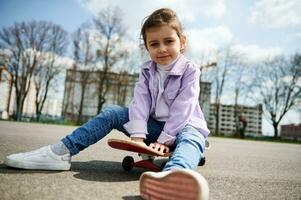 Cute female child posing to the camera while having rest sitting on a wooden skateboard on the playground photo