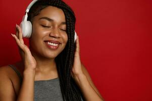 Stunning beautiful African woman smiles toothy smile, posing with closed eyes from enjoyment and pleasure while listening to the music in wireless headphones, against red background with copy space photo