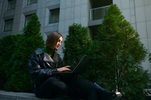 Successful beautiful woman, business person, freelancer, entrepreneur working on laptop on the background modern office corporate buildings. photo