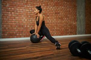 Charming young African athlete in a stylish tight-fitting tracksuit performing side lunges, leaning her hands on a medicine ball photo