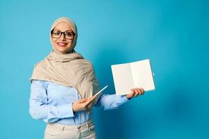 Smiling Muslim woman in hijab holds blank book and points at it with pen isolated on blue background photo