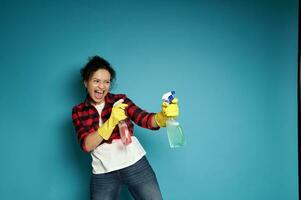 Young woman playing with cleaning sprays as if shooting a gun. Posing over blue background with copy space photo