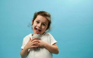 Cute girl gently hugs her soft toy bear and looks at the camera, standing on a blue background with copy space photo