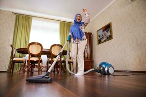 Cheerful charming young Arab Muslim woman, housewife, housekeeper with head covered in blue hijab vacuuming floor, dancing and singing while cleaning house photo