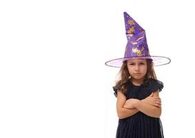 Portrait of cute little witch angry upset girl wearing a wizard hat and dressed in stylish carnival dress, looking at camera posing with crossed arms against white background, copy space, Halloween photo