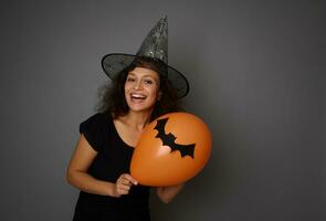 Beautiful young woman in black and wizard hat poses with an orange air balloon with a handmade felt-cut bat, smiles toothy smile looking at camera. Halloween concept on grey background, copy space photo