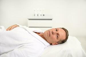 Handsome middle aged attractive Caucasian man in white terry robe looks at the camera lying down on a massage table in white luxury interior of a massage room in wellness spa center photo