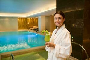 Charming woman in white bathrobe, holds a glass with delicious healthy freshly squeezed vitamin juice in her hands, smiles looking at camera, standing by the jacuzzi at wellness spa centre lounge area photo