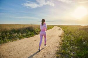 posterior ver de joven ajuste mujer en rosado ropa de deporte corriendo en naturaleza con hermosa luz de sol foto