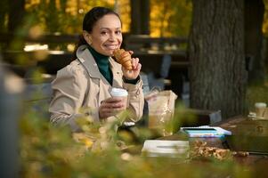 confidente negocio mujer disfruta su desayuno, come cuerno y bebidas café , resfriado lejos desde el ajetreo y bullicio de el ciudad, sonrisas mirando a cámara antes de distante trabajo en acogedor al aire libre café foto