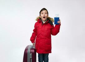 Happy school child, adorable handsome boy in bright red down jacket standing near a suitcase and holding his passport with ticket and boarding pass. Travel and winter holidays concept with copy space photo