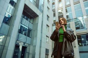 Business and communications concept. Beautiful self-confident European young woman, freelancer, entrepreneur, copywriter using mobile phone on modern office corporate high-rise buildings background. photo