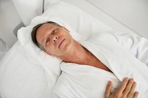 High angle view of handsome relaxed mature 50 years old Caucasian man in white terry bathrobe looking at camera while lying on massage table in white luxury wellness spa clinic photo