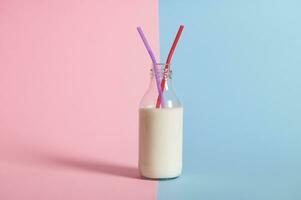 Closeup of transparent bottle with milk and two straws in it, isolated on bicolor blue and pink background photo