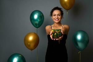 elegante Hispano mujer en negro noche vestir muestra un Navidad presente en brillante envase papel y dorado arco, sonrisas con dientes sonrisa mirando a cámara. gris antecedentes con oro verde metálico globos foto