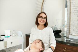 sonriente cosmetóloga sentado cerca un cliente mujer en contemporáneo belleza spa clínica. foto