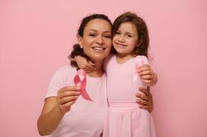 African American woman hugs her daughter, holding pink ribbon, cute smiles looking at camera, isolated on colored background with copy space. International Day of fight against a Breast Cancer disease photo