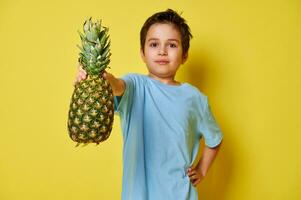 Handsome child boy holding a pineapple while posing over yellow background. Summer and healthy vegan food concept photo