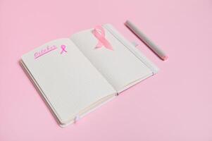 Lettering 1 st October on a diary and a pink ribbon on an empty blank paper sheet, isolated on pink background with copy space. World Breast Cancer Day photo